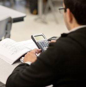 A student is seen from behind, using a calculator and referring to a math text book at a desk