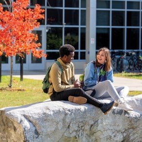 Two students sitting on a large rock outside of the residence hall