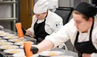 Two Culinary students dressed in chefs' uniforms and hats plate food at an event at Seasoned.
