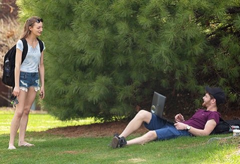Two students visit, while one lounges in the grass with a laptop and the other stands with a backpack
