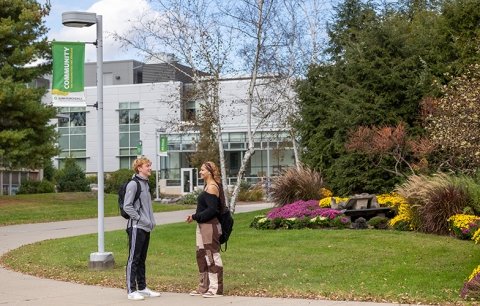 Two students are talking in front of Adirondack Hall with fall foliage in the background