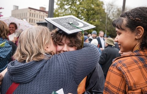 A graduate in cap and gown is seen over the shoulder of a loved one they are hugging